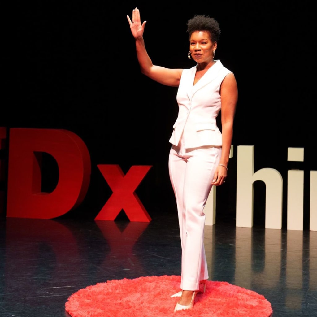 A woman in a white suit stands on a red circular carpet, raising one hand while speaking as a personal stylist on stage with TEDx signage in the background. Personal stylist Los Angeles